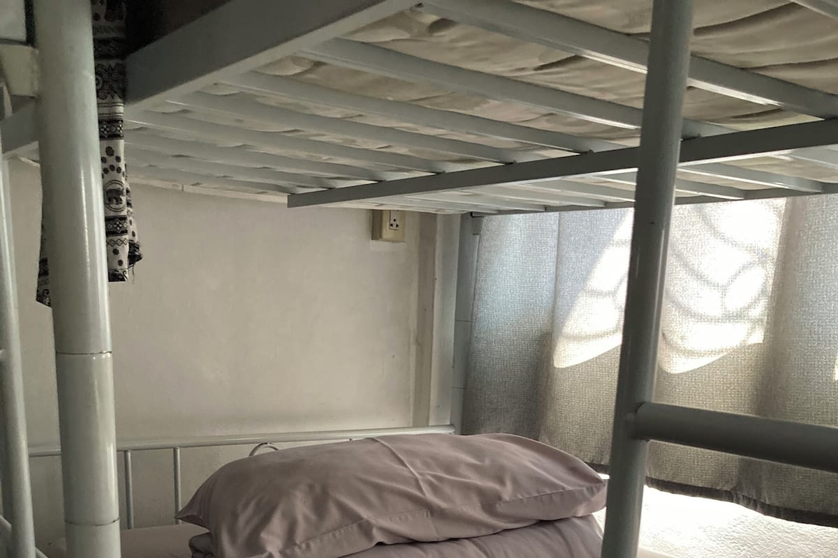 Un lit superposé avec un matelas simple et un oreiller, une fenêtre avec des rideaux légers et une prise électrique au mur.