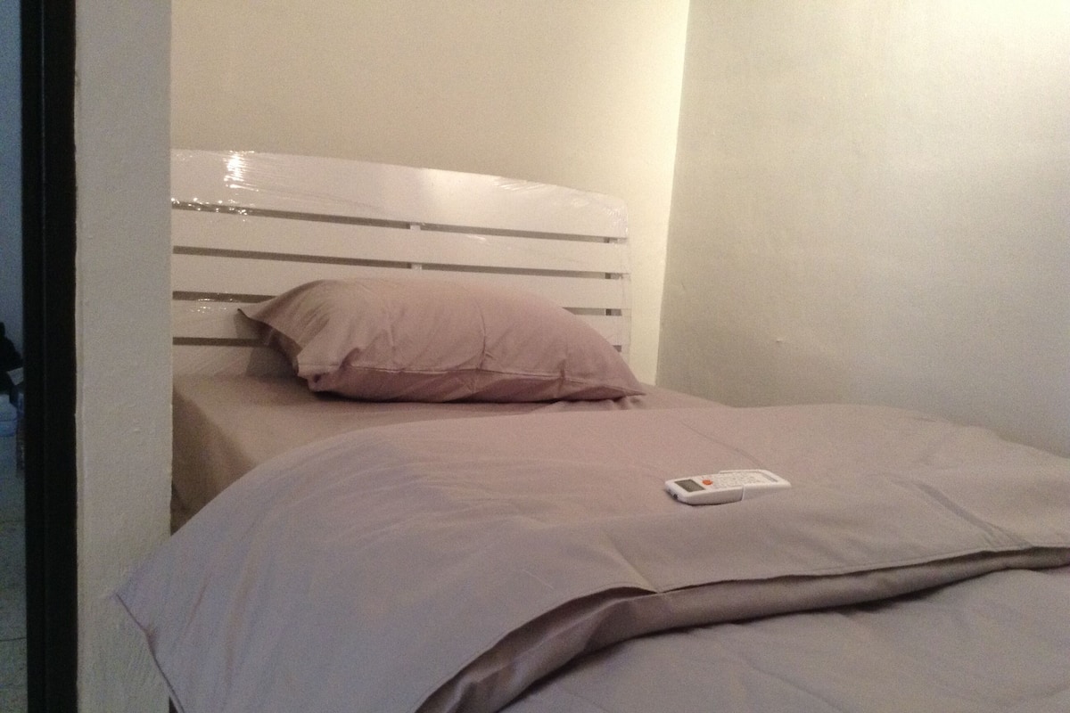 Un lit simple avec une tête de lit blanche, un oreiller rose et une télécommande posée sur le lit.