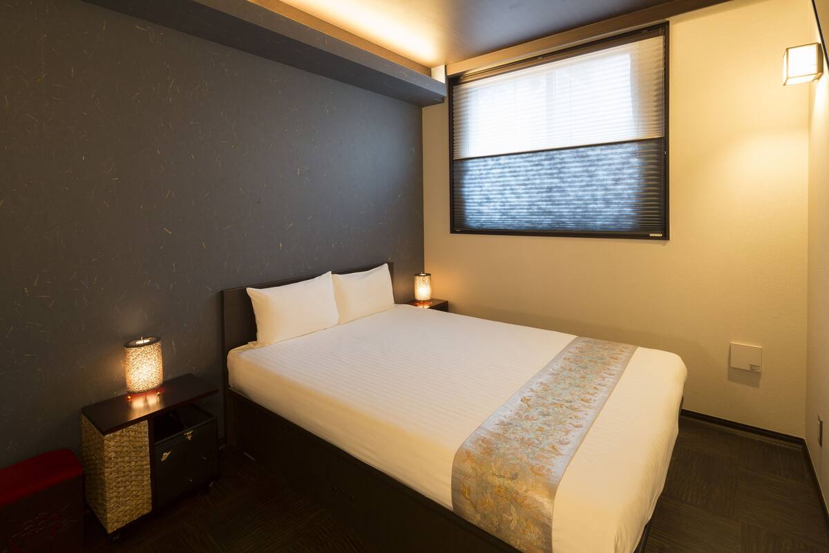 A neatly made bed with a white comforter and a decorative border, a bedside table with a lamp, and a window with blinds.