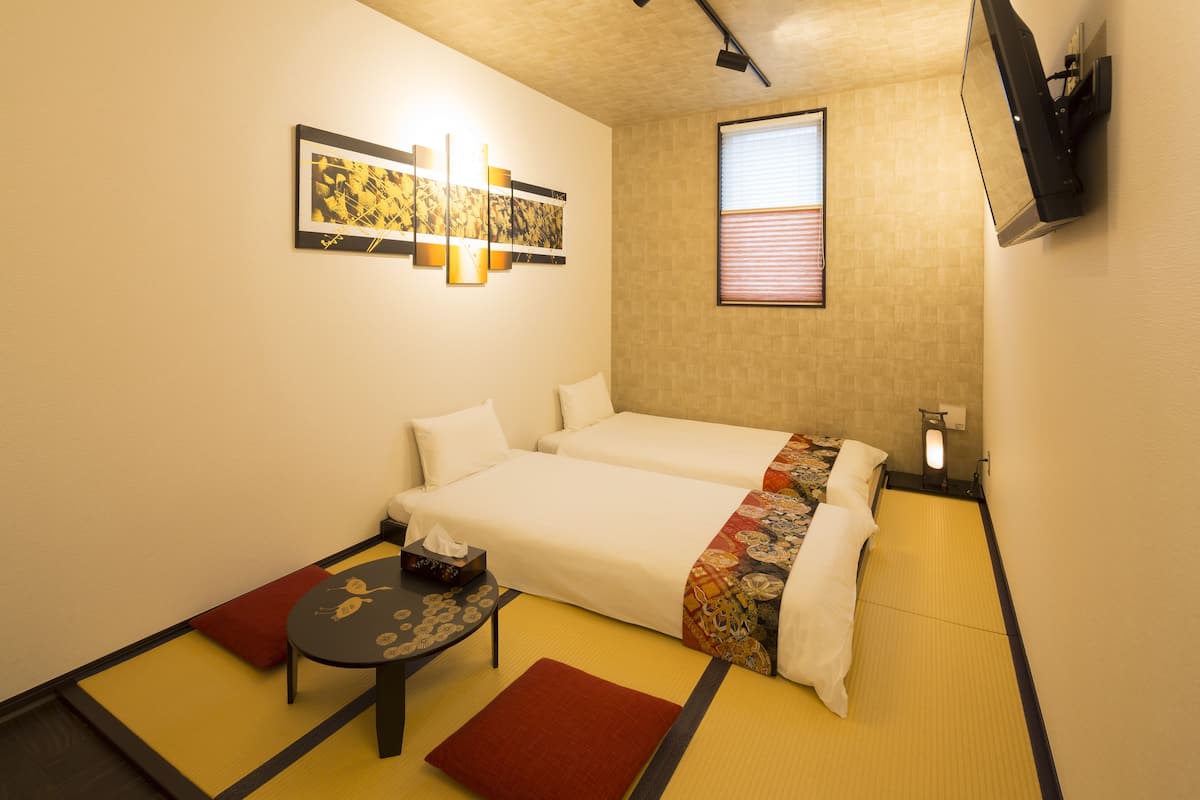 A hotel room with two beds, a small table, a TV mounted on the wall, and a window with blinds.