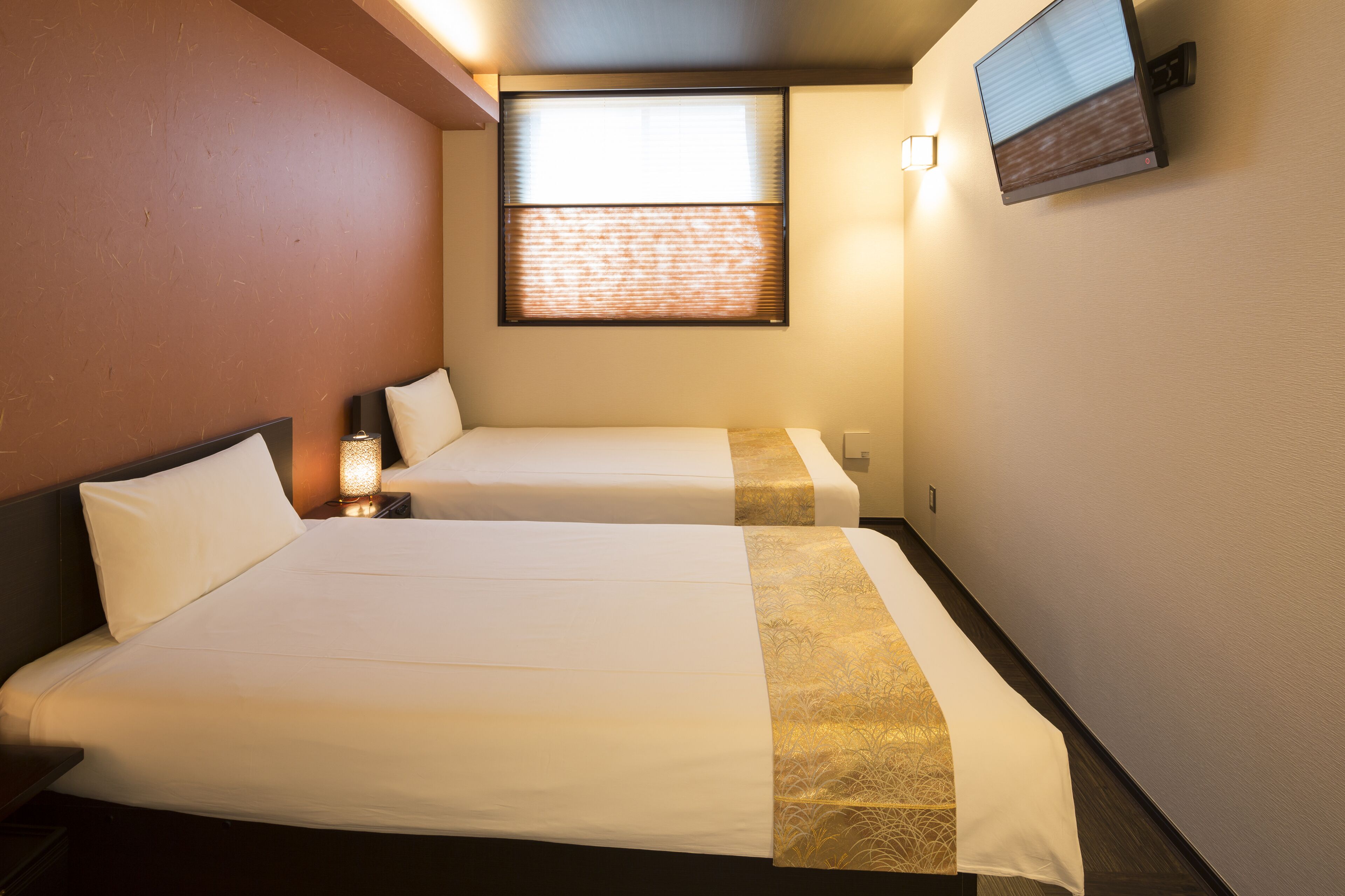 A hotel room with two beds, a window with blinds, a flat-screen TV mounted on the wall, and a nightstand with a lamp.