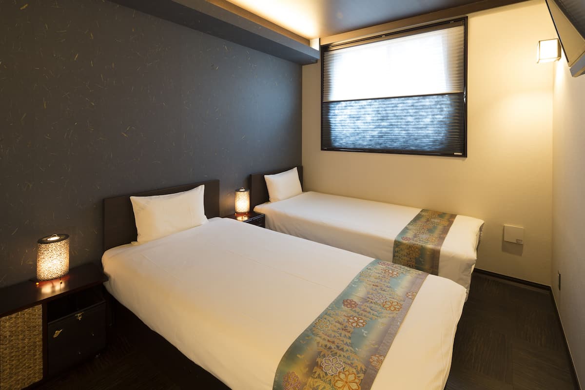 A hotel room with two beds, a nightstand with a lamp, a window with blinds, and a wall-mounted light switch.