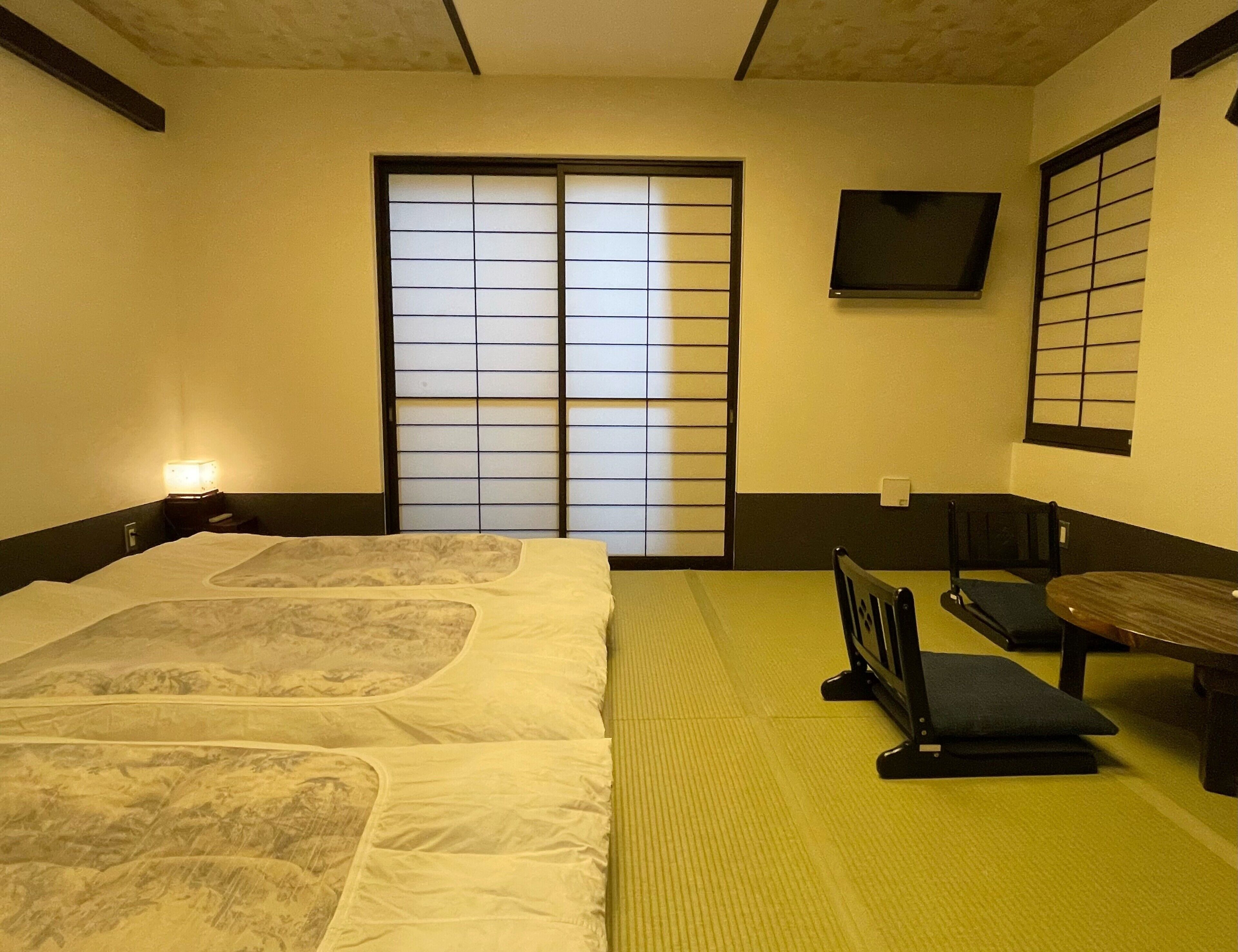 A traditional Japanese room with tatami flooring, a futon bed, sliding shoji doors, and a television mounted on the wall.