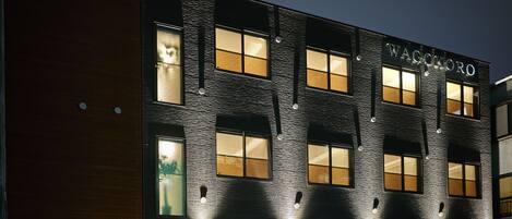A building with the name "WAGOKORO" illuminated at night, featuring a modern architectural design with multiple windows and a green fence in the foreground.