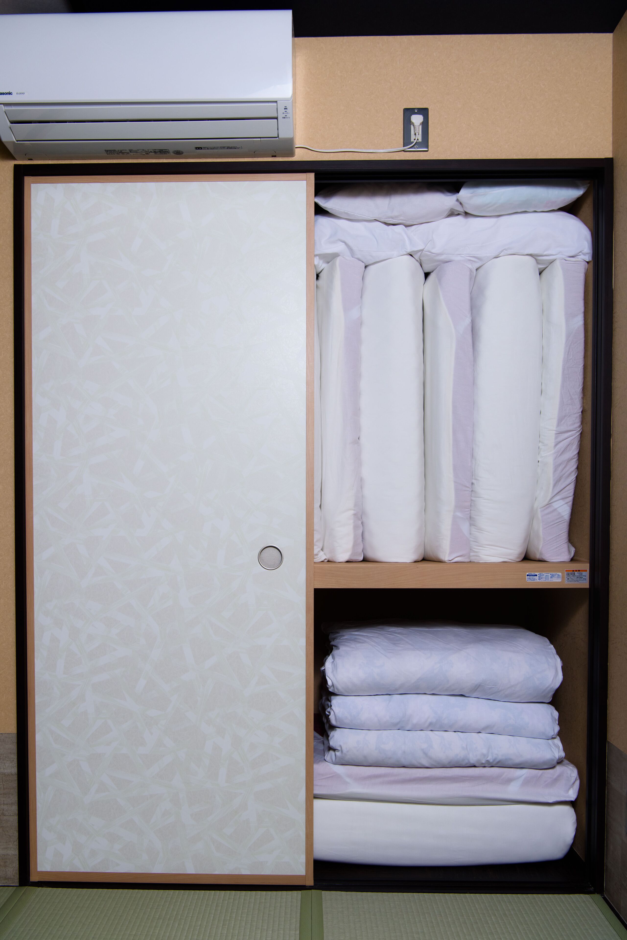 A hotel room with a large air conditioning unit, a wardrobe with a patterned door, and neatly stacked pillows and blankets.