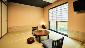 Una habitación con tatamis, una mesa baja, sillas, un celosía japonesa y un televisor montado en la pared.