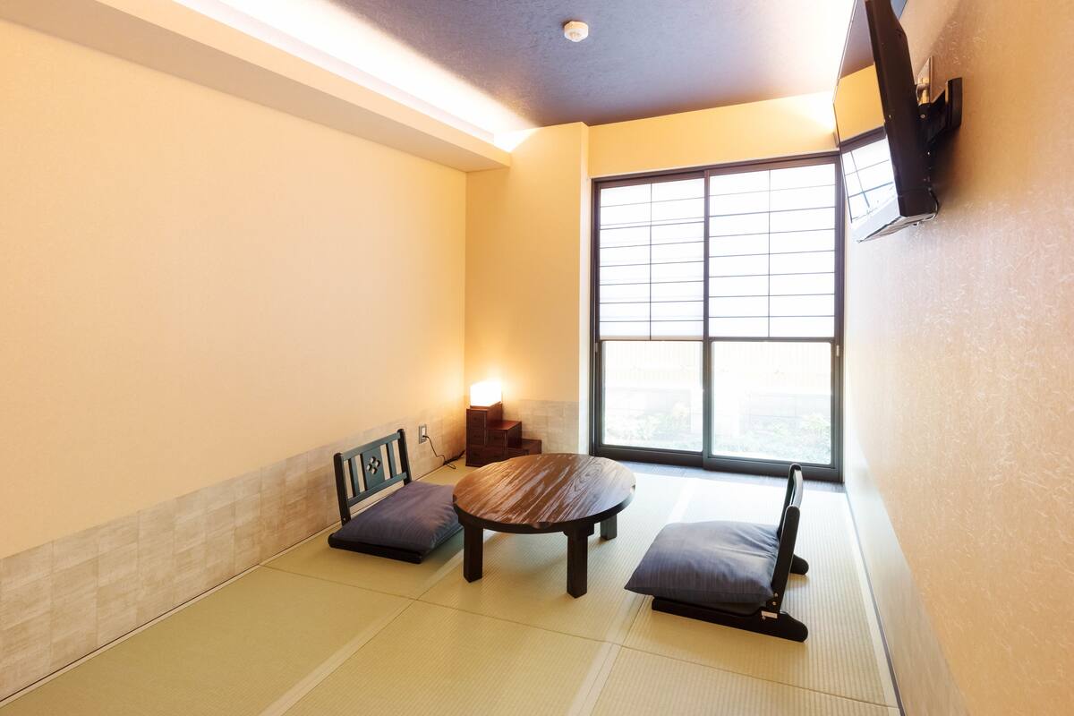 A tatami mat room with a round table, two cushions, a low wooden bench, a wall-mounted TV, and a window with a view.