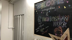Un aula con una pizarra que muestra 'WELCOME TO TOKAGOSHIMA & TENMONKAN PARK' en inglés y japonés, una pizarra blanca y una puerta con cortina.