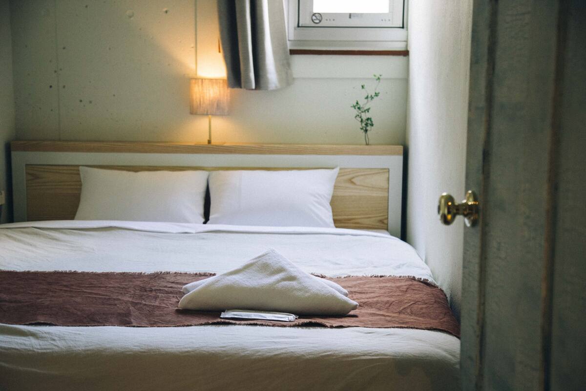 Una cama bien hecha con sábanas blancas y una manta marrón, una lámpara de noche y una planta en la pared.