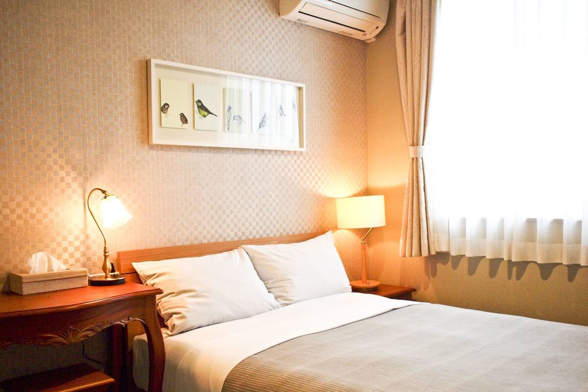 Ein Hotelzimmer mit einem Bett, zwei Nachttischlampen, einem Nachttisch, einer Taschentuchbox und einem an der Wand hängenden Bild von Vögeln.