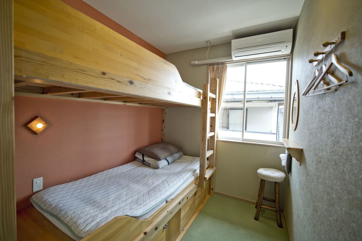 Ein kleines Zimmer mit einem Etagenbett, einem kleinen Hocker, einem Fenster mit Vorhängen und einer Klimaanlage.