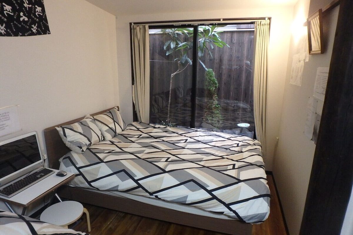 Een slaapkamer met een bed, een laptop op een klein tafeltje, en uitzicht op een houten schutting en beplanting.