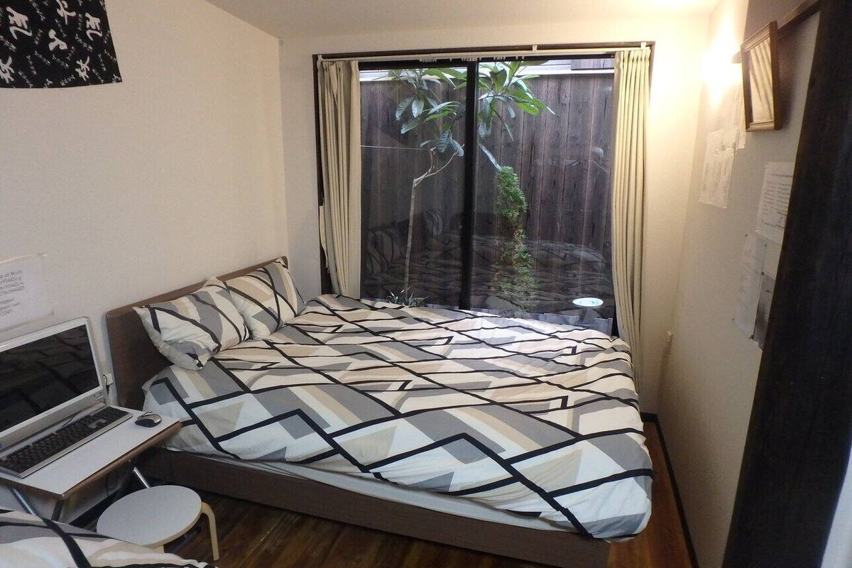 Een slaapkamer met een bed, een laptop op een klein tafeltje, en uitzicht op een houten schutting en beplanting.