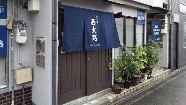 Un bâtiment doté d’un auvent bleu orné de caractères japonais, d’une porte marron et de plantes en pot disposées sur le trottoir.