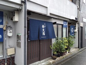 Un edificio con toldo azul que muestra caracteres japoneses, una reja marrón y macetas con plantas en la acera.