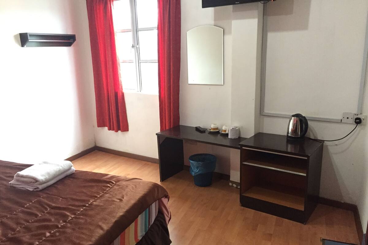 Une chambre d’hôtel avec un lit, un bureau, une chaise, une poubelle et une fenêtre avec des rideaux rouges.