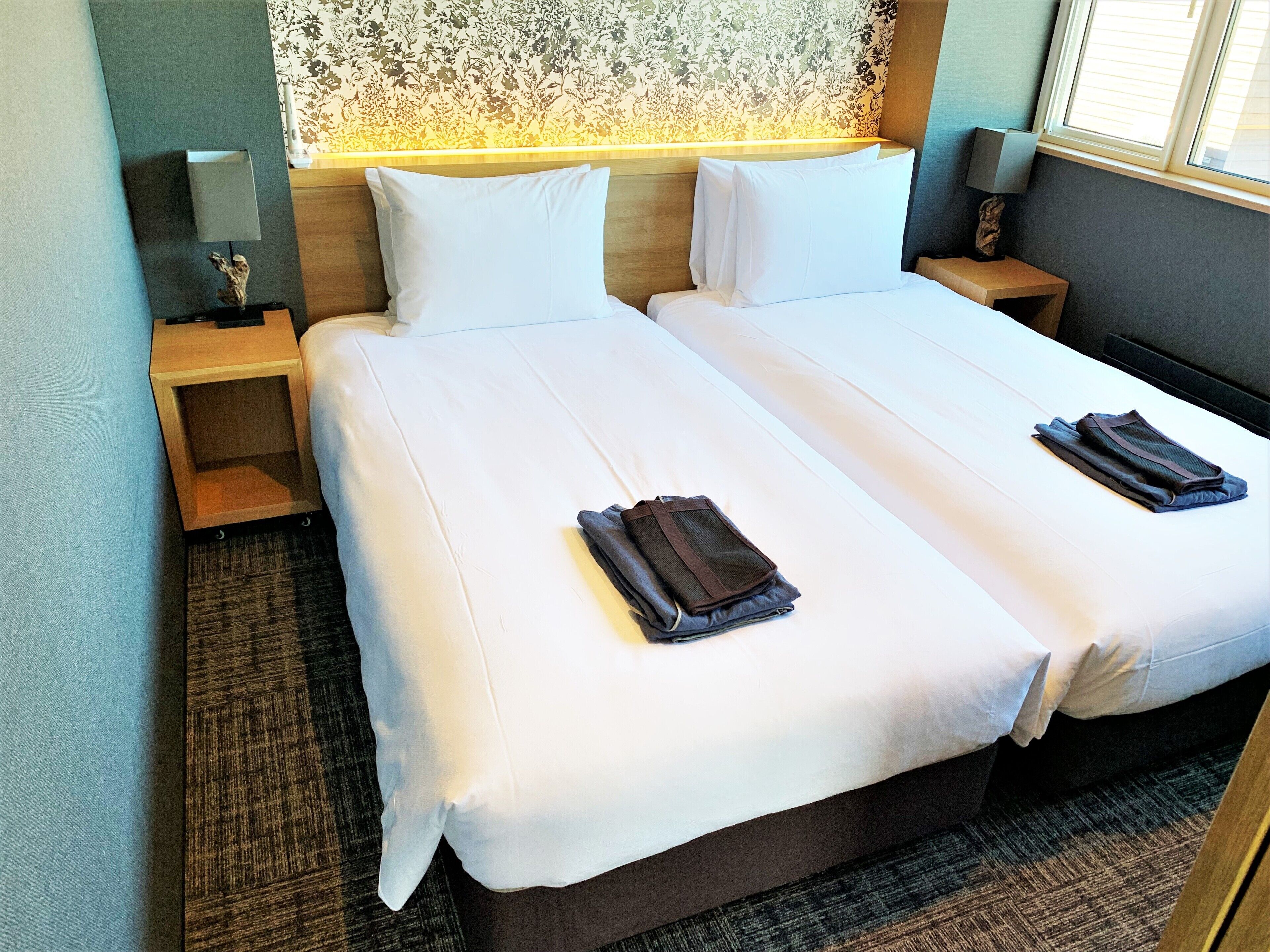 A double bed with white linens and two folded towels. There is a nightstand with a lamp and a small statue. The room has a floral wallpaper and a window with blinds.