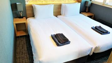 A double bed with white linens and two folded towels. There is a nightstand with a lamp and a small statue. The room has a floral wallpaper and a window with blinds.