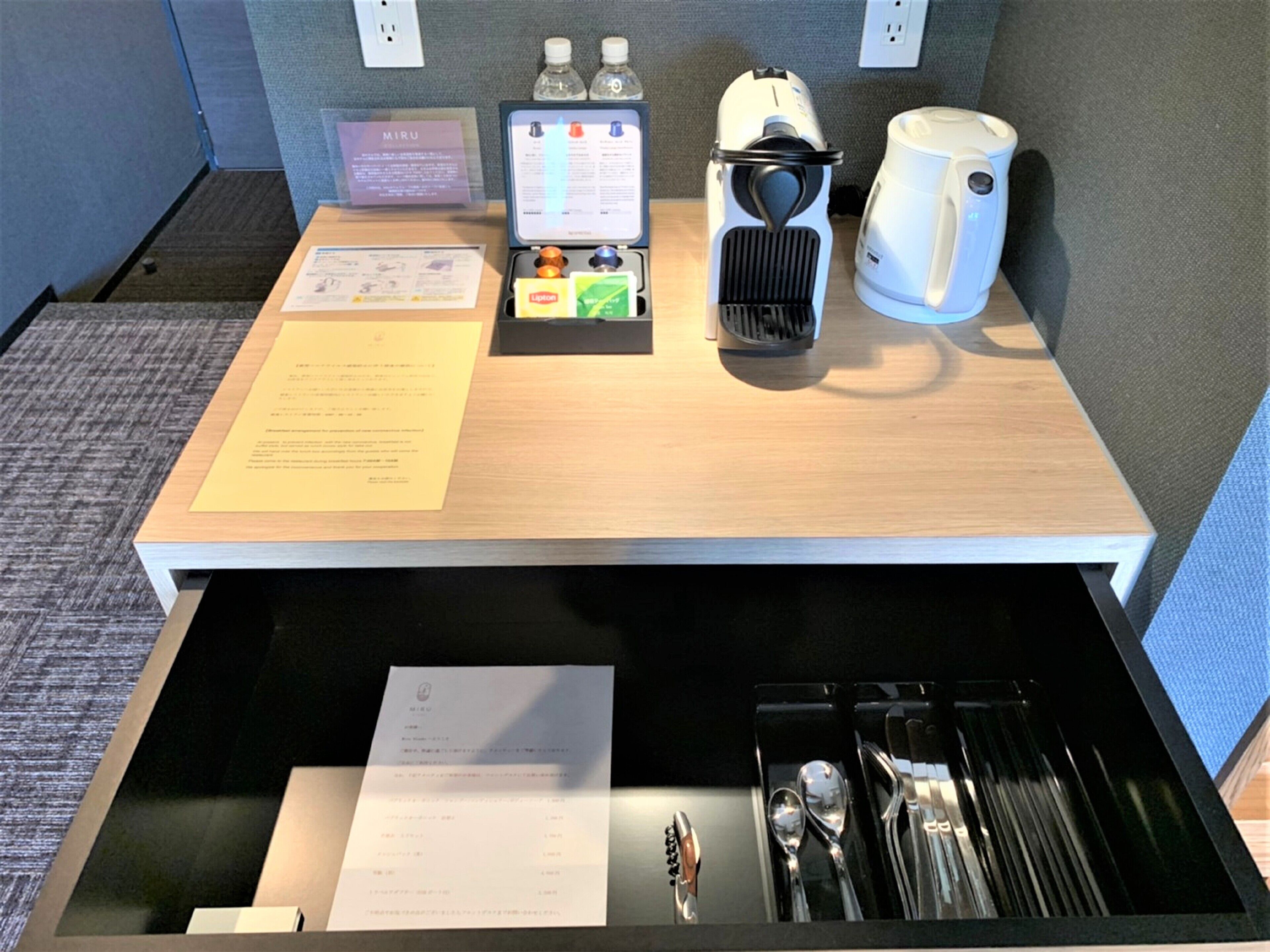 Habitación de hotel con escritorio de madera, cafetera, botella de agua, mini refrigerador y un cajón con cubiertos.