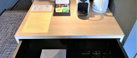 Habitación de hotel con escritorio de madera, cafetera, botella de agua, mini refrigerador y un cajón con cubiertos.