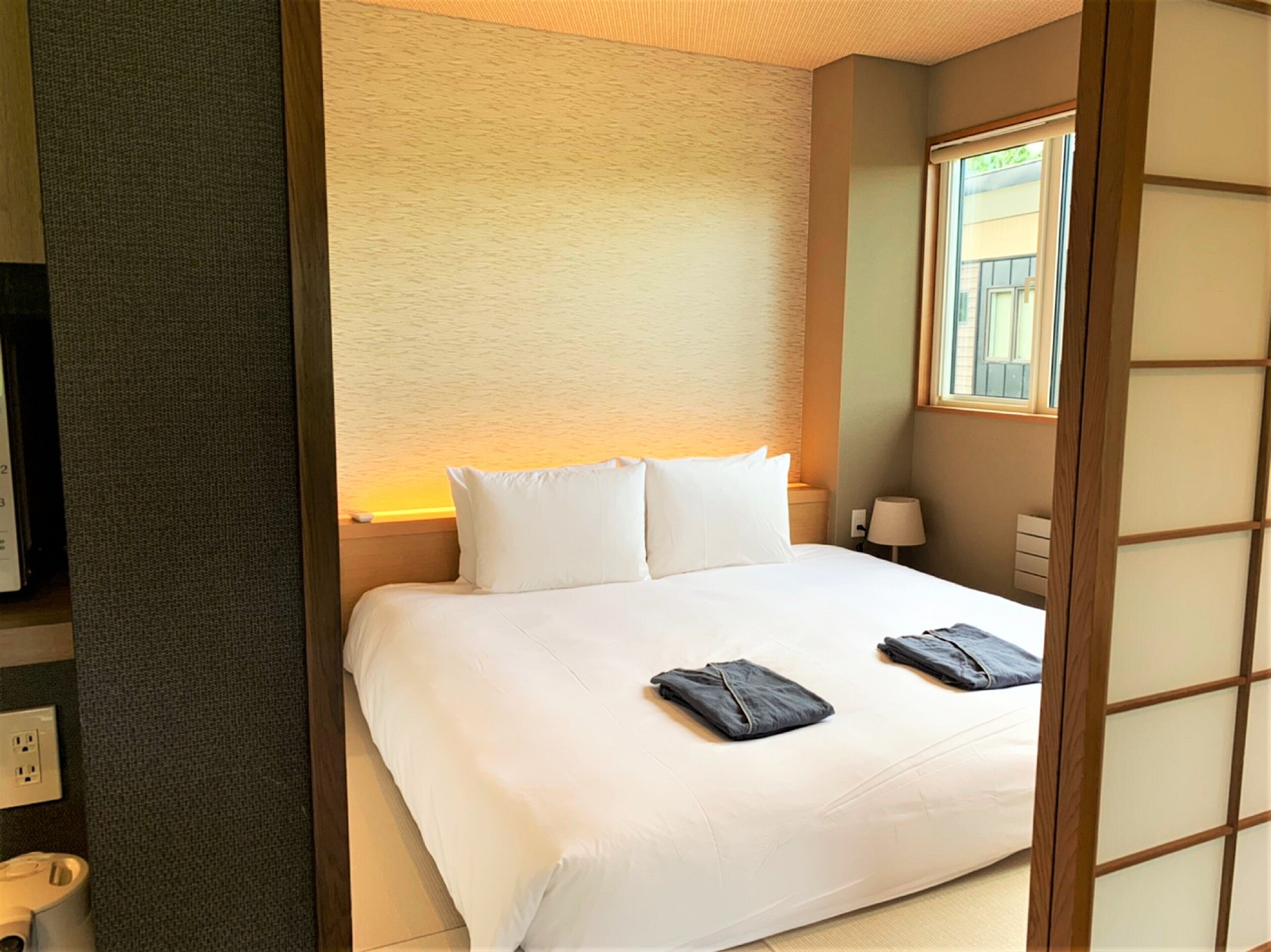 Una habitación de hotel con una cama, una mesita de noche, una lámpara y una ventana.