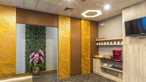 Un moderno vestíbulo de hotel con un mostrador de recepción, una pared verde con plantas, una máquina de café y un televisor en la pared.
