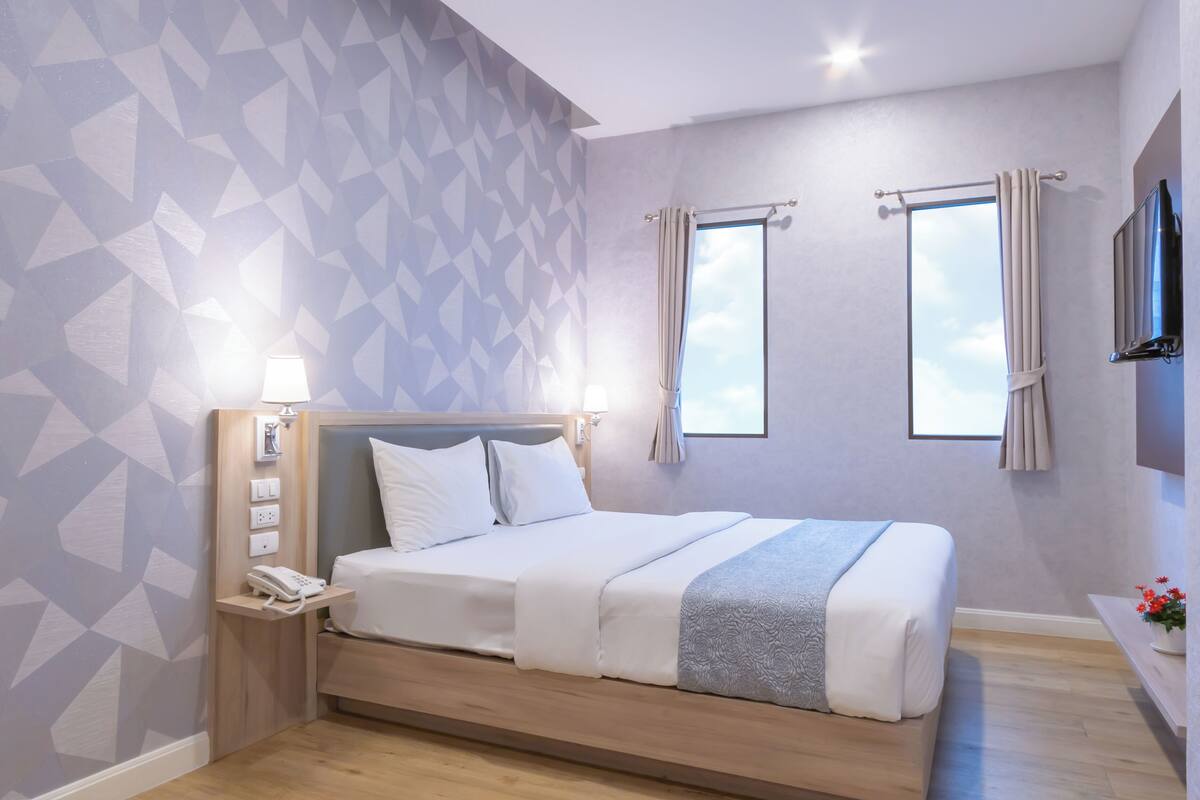 Une chambre d’hôtel moderne avec un mur orné d’un motif géométrique, un lit en bois avec une literie blanche, un téléphone sur la table de chevet et une fenêtre avec des rideaux.