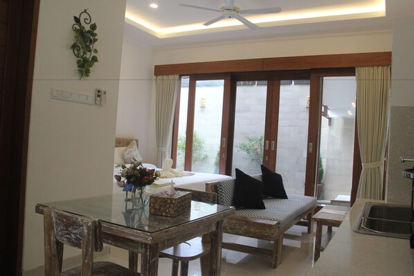 Habitación de hotel con mesa de comedor de vidrio, sillas de madera, sofá con cojines negros, ventilador de techo y vistas a un espacio exterior a través de grandes puertas correderas.