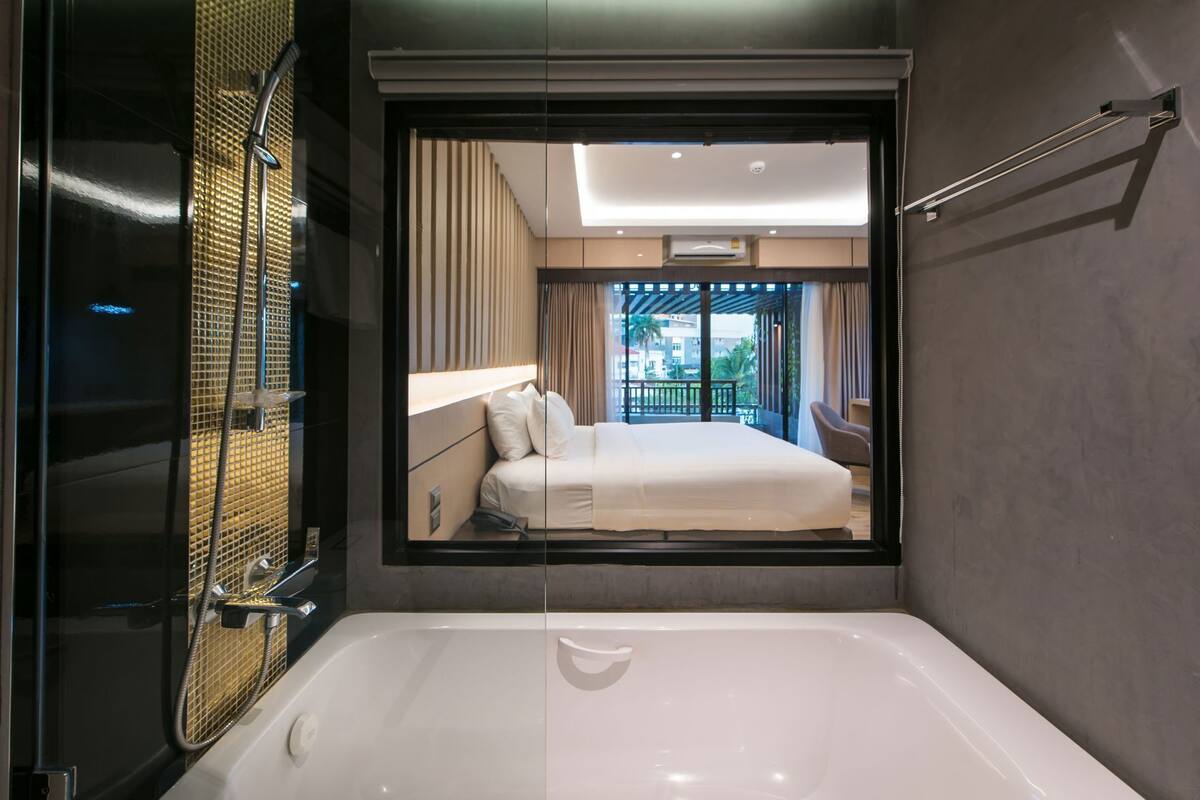 A modern bathroom with a large bathtub, a shower area, and a view of a bedroom with a bed and curtains.