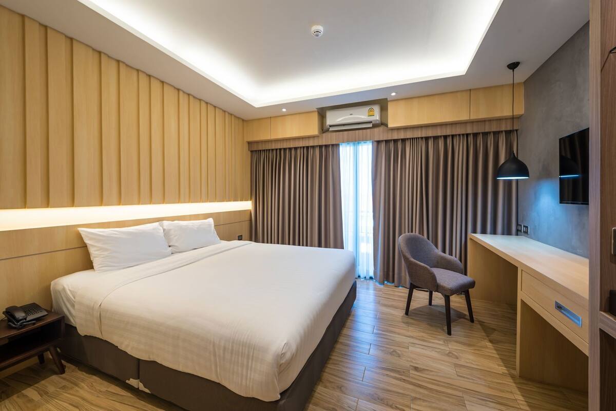 A hotel room with a bed, a desk, a chair, an air conditioning unit, and a window with curtains.