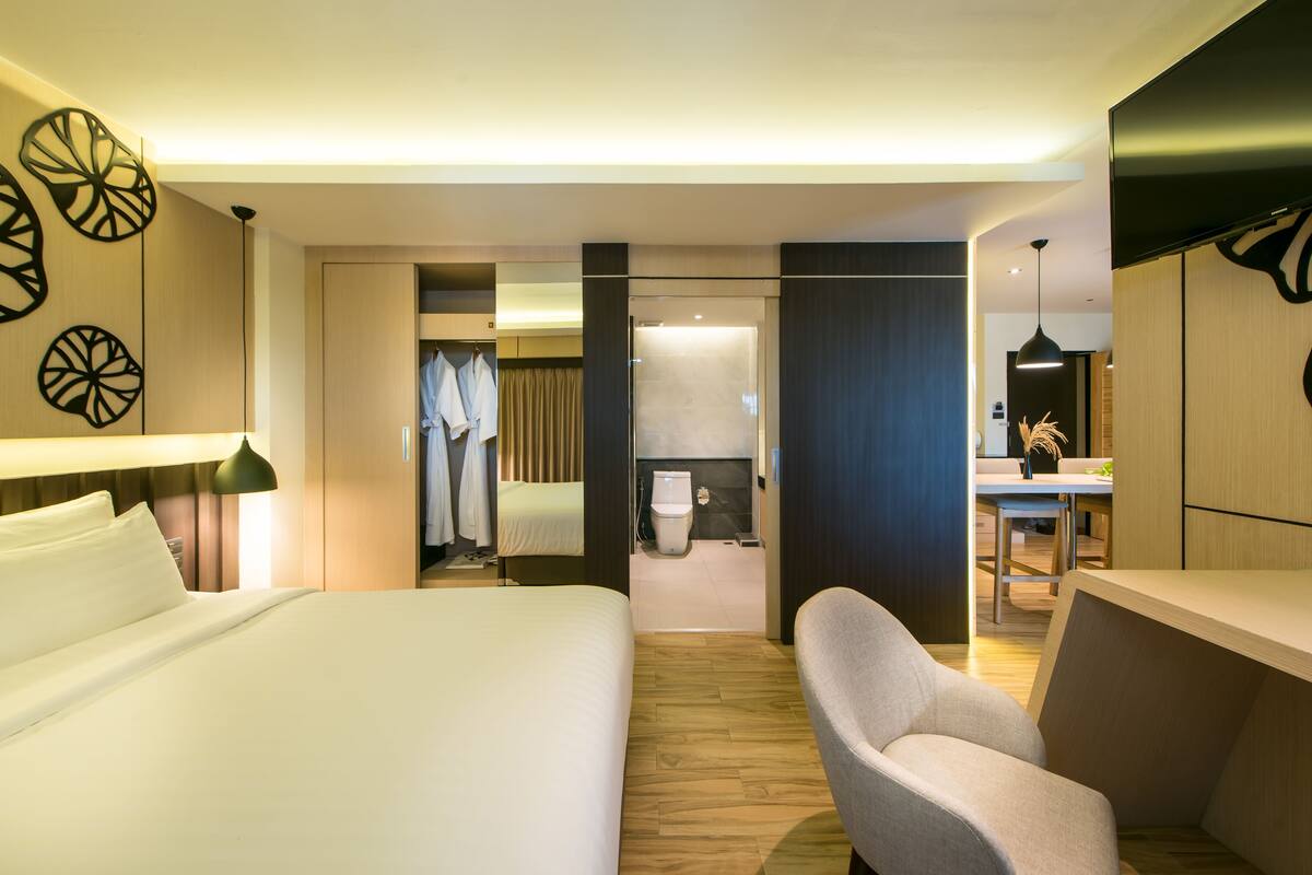 A hotel room with a large bed, a desk, a chair, a wardrobe, and a bathroom visible through an open door.