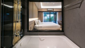 A modern bathroom with a large bathtub, a shower area, and a large window providing a view of the room and outside. - Nadee 10 Resort & Hotel (Khon Kaen)