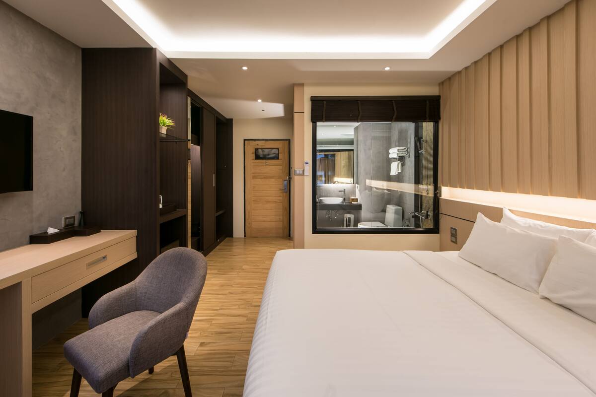A modern hotel room with a wooden desk, a comfortable chair, a large bed, and a bathroom visible through a glass partition.