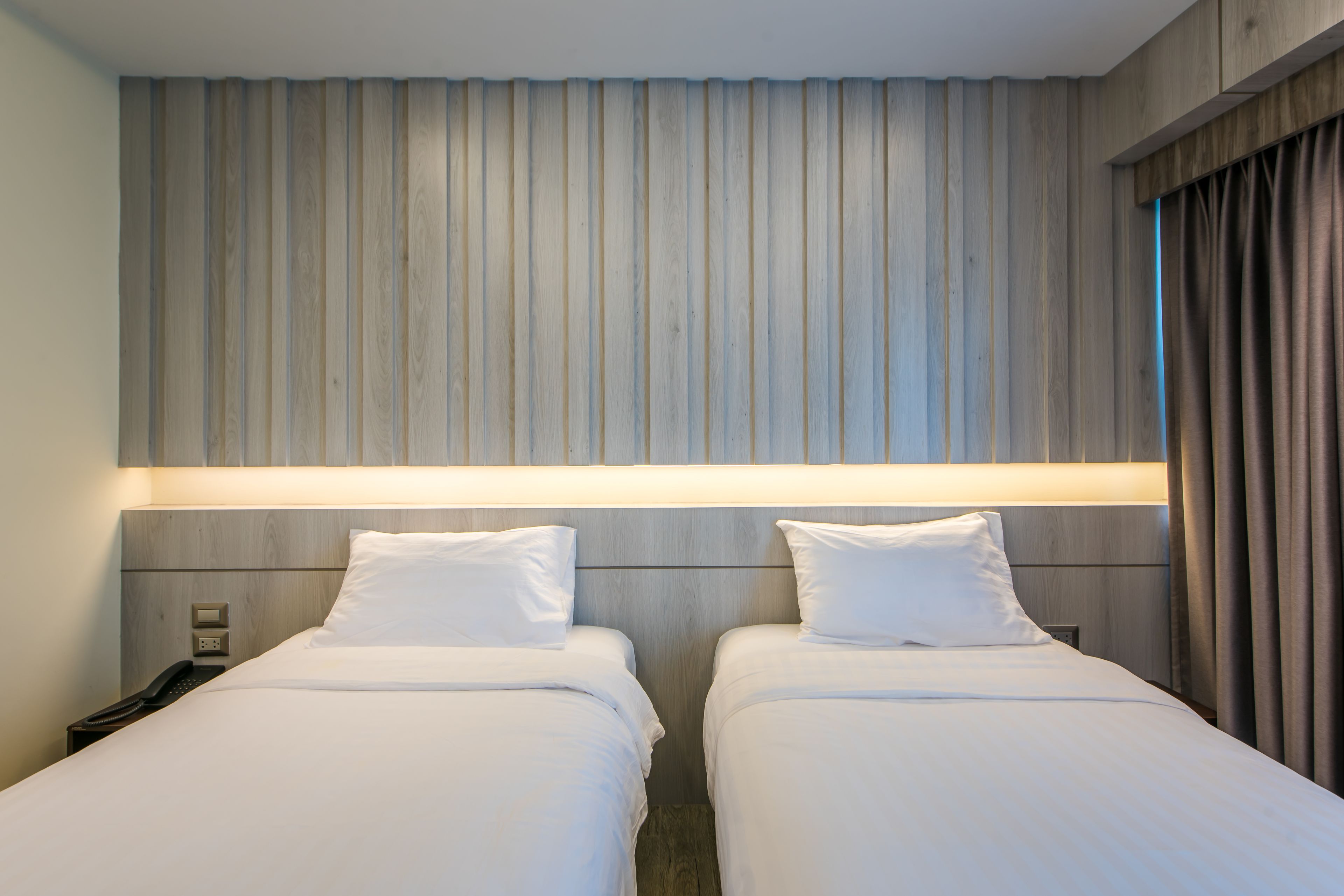 A hotel room with two beds, a wooden headboard, a bedside table with a phone, and a window with curtains.