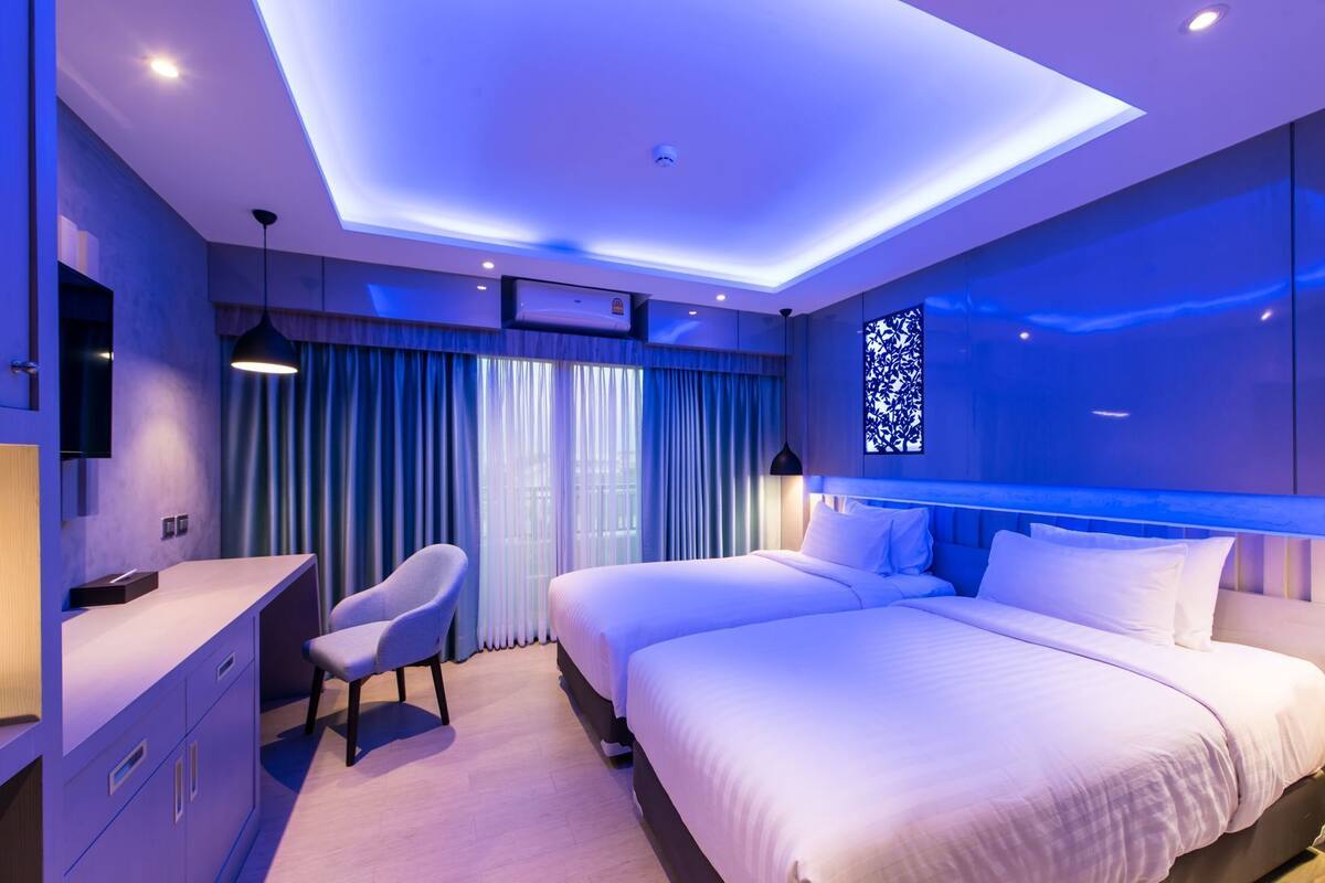 A modern hotel room with a desk, two beds, a chair, and a large window with curtains.