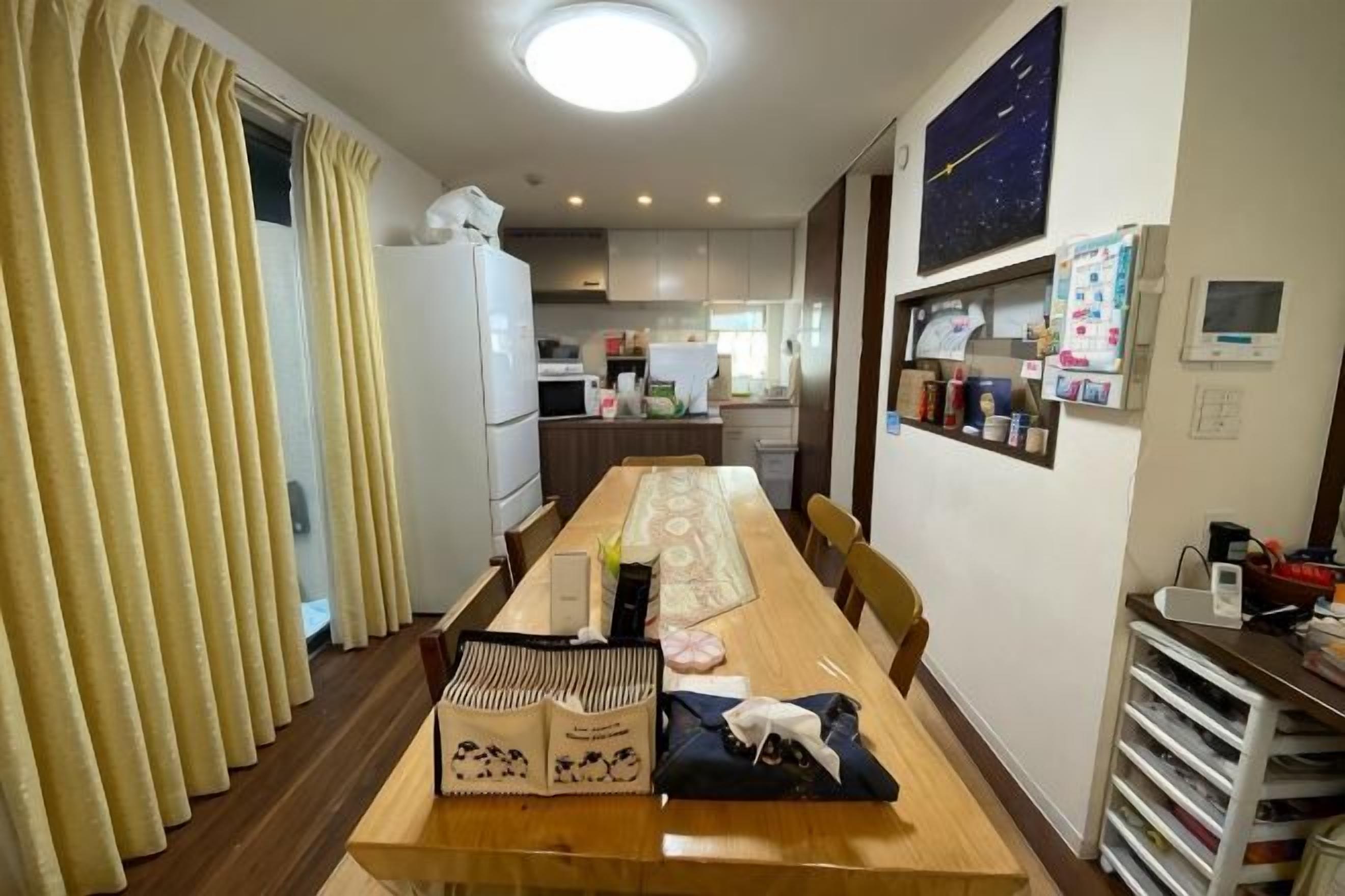 A dining area with a long table, chairs, a refrigerator, and a kitchen in the background.