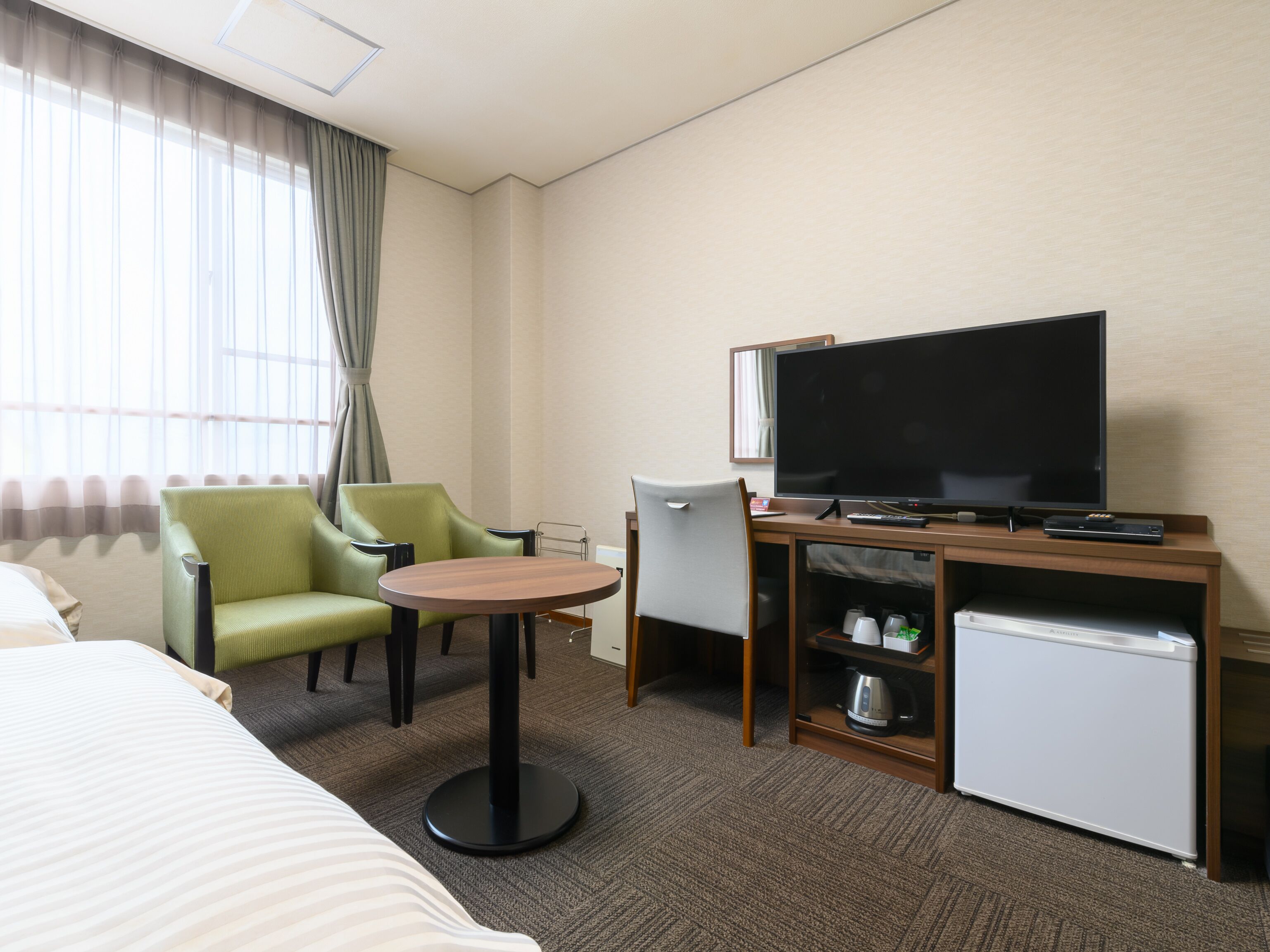 Una habitación de hotel con cama, escritorio, televisión, refrigerador pequeño y una silla.