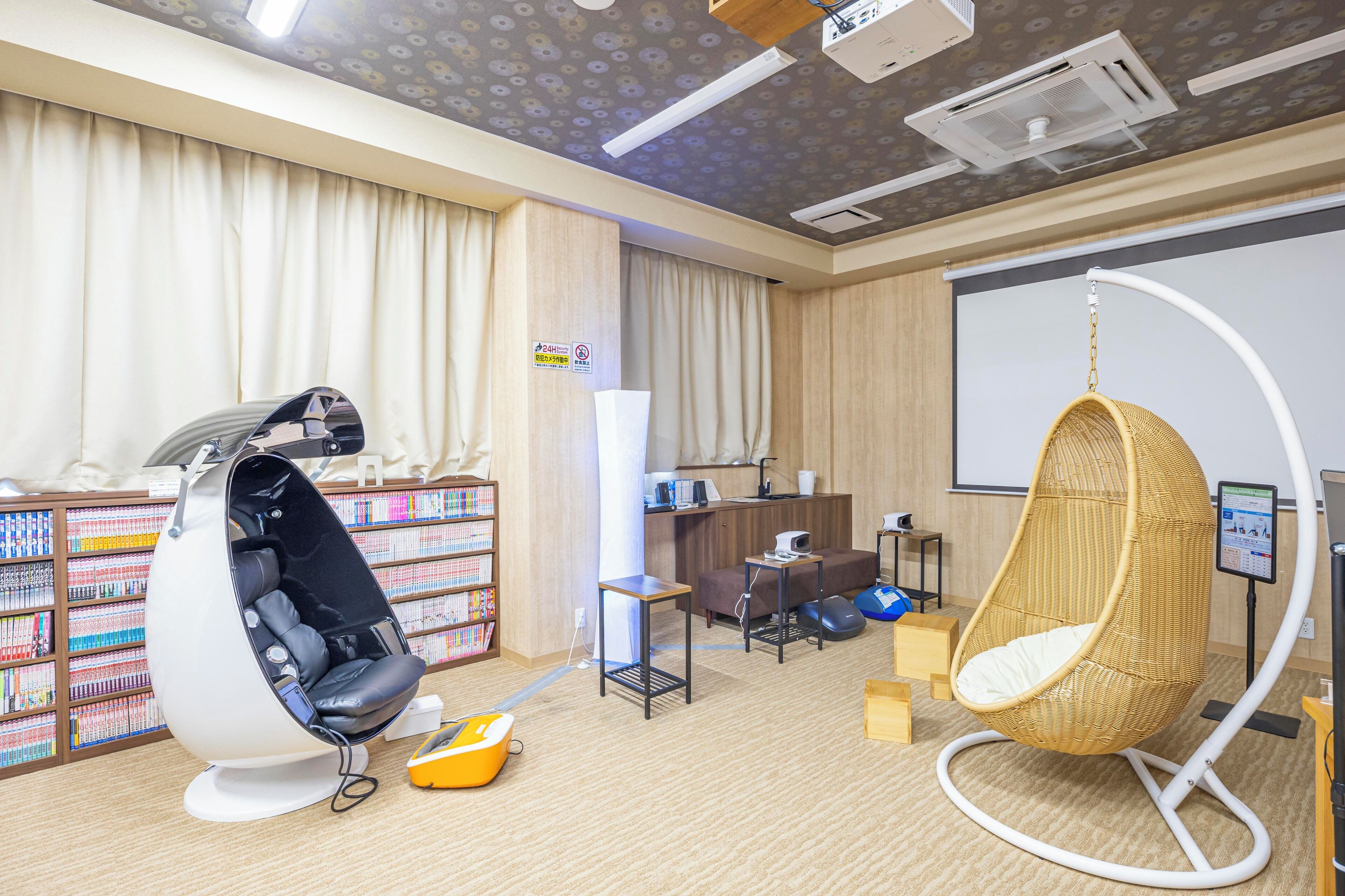 Ein Zimmer mit einem weißen Ei-Sessel, einem Hängesessel aus Weidengeflecht, einem Bücherregal, einem Schreibtisch und einer Projektionsleinwand.
