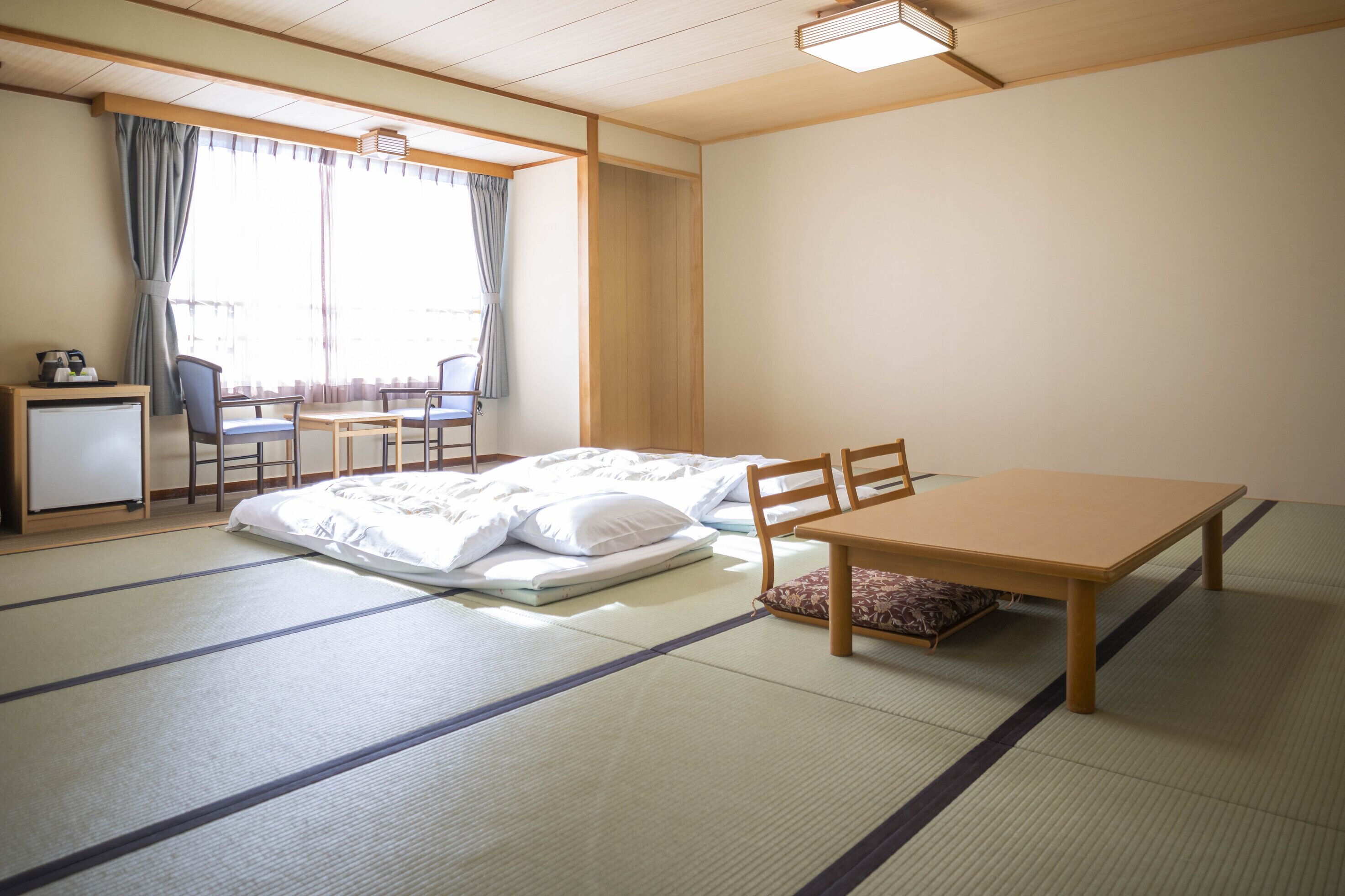 A room with tatami flooring, a bed with white bedding, a wooden table, a chair, a desk with a chair, and a window with curtains.