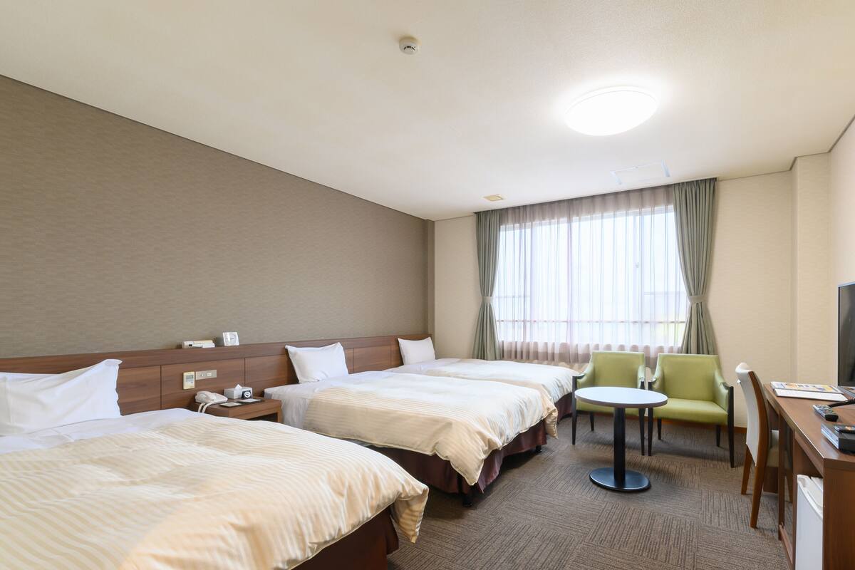 A hotel room with two beds, a small round table, a chair, a desk with a TV, and a window with curtains.