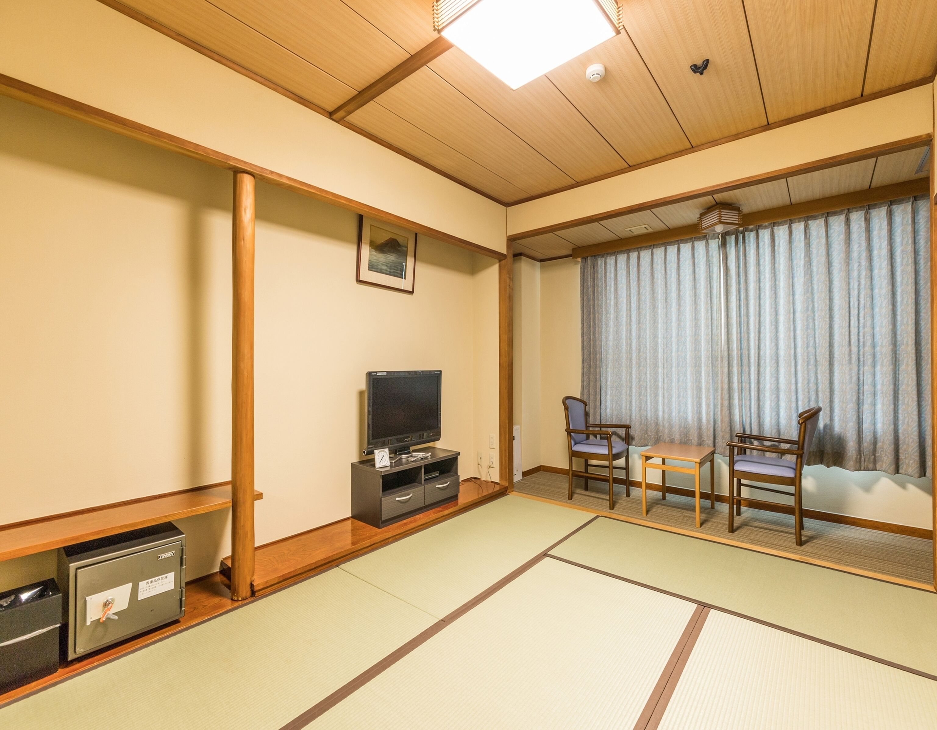 A traditional Japanese room with tatami flooring, a television on a low stand, a small table, two chairs, and a sliding door with curtains.