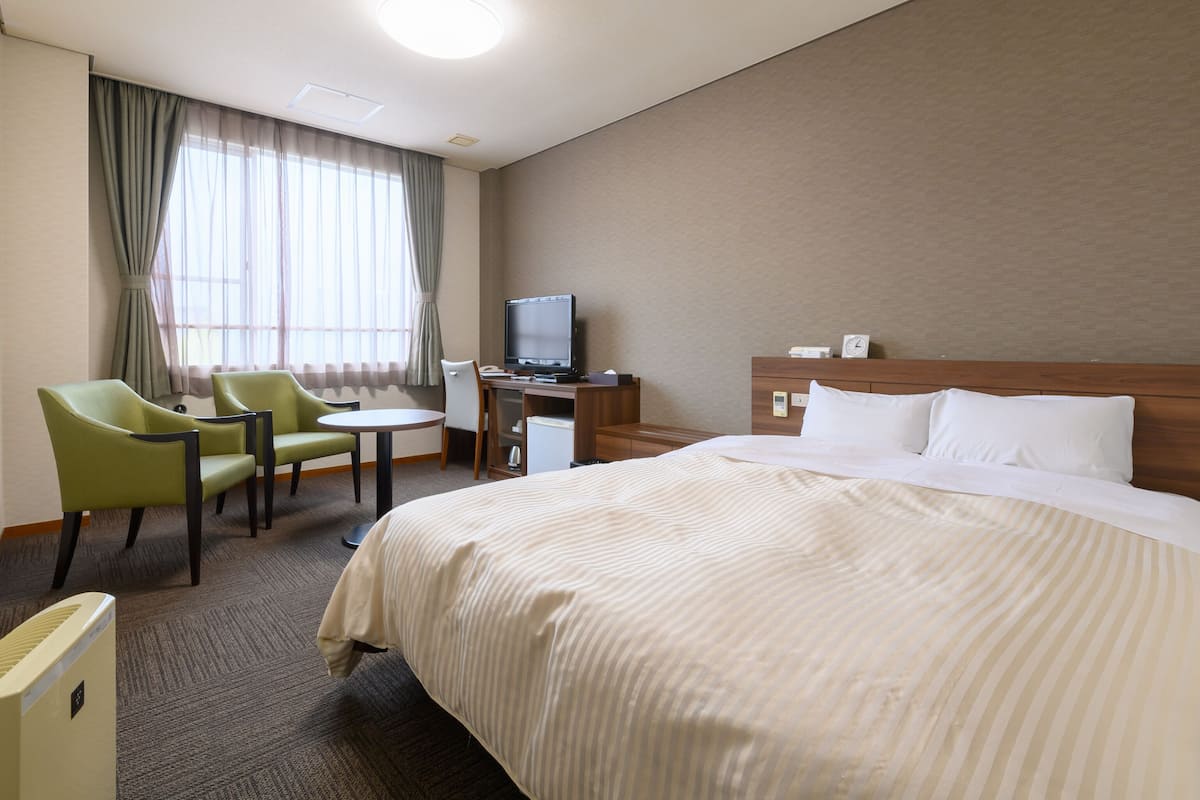 A hotel room with a bed, a desk with a computer, two chairs, a television, and a window with curtains.