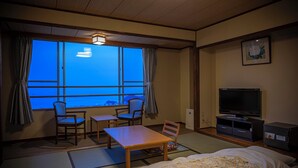 Une chambre d’hôtel avec un sol recouvert de tatamis, un lit, une télévision, un bureau, deux chaises et une fenêtre avec des rideaux.