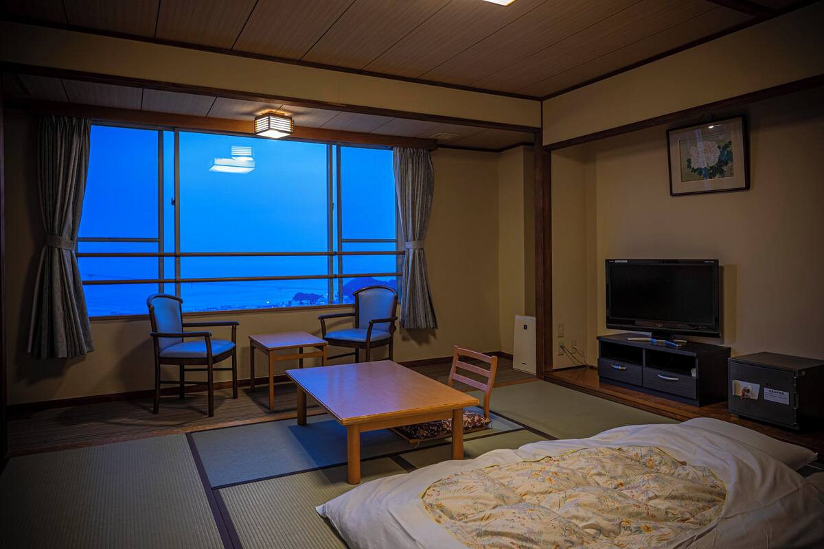 A hotel room with tatami flooring, a bed, a television, a desk, two chairs, and a window with curtains.