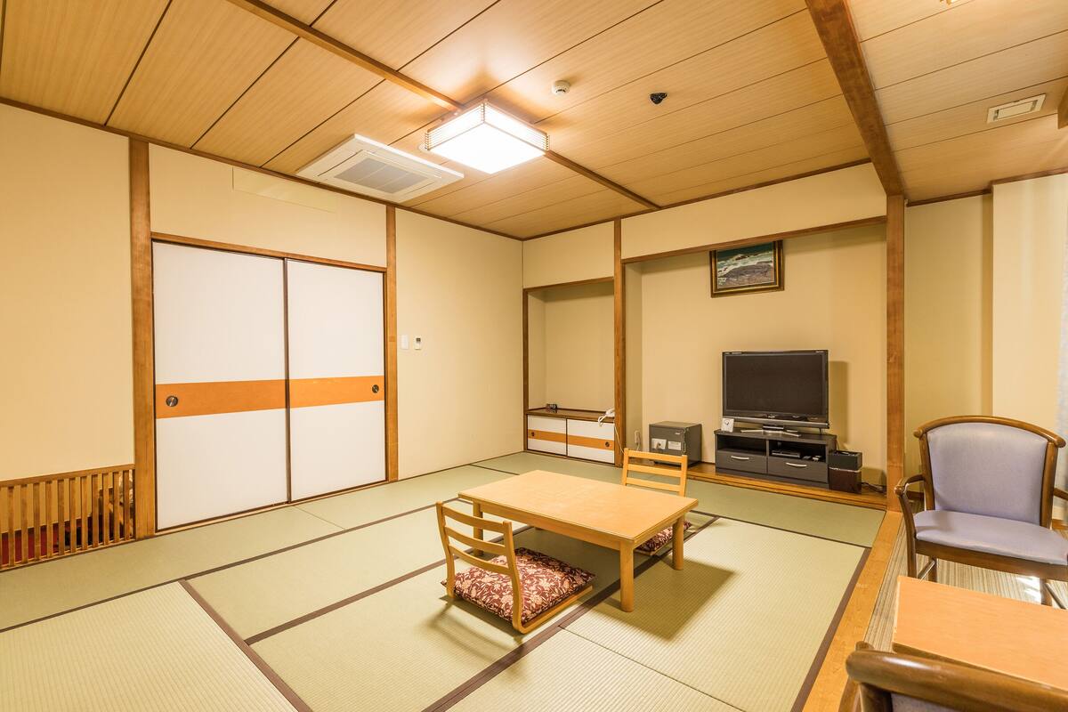 A traditional Japanese room with tatami flooring, a low table with a cushion, a television, and a wooden chair.
