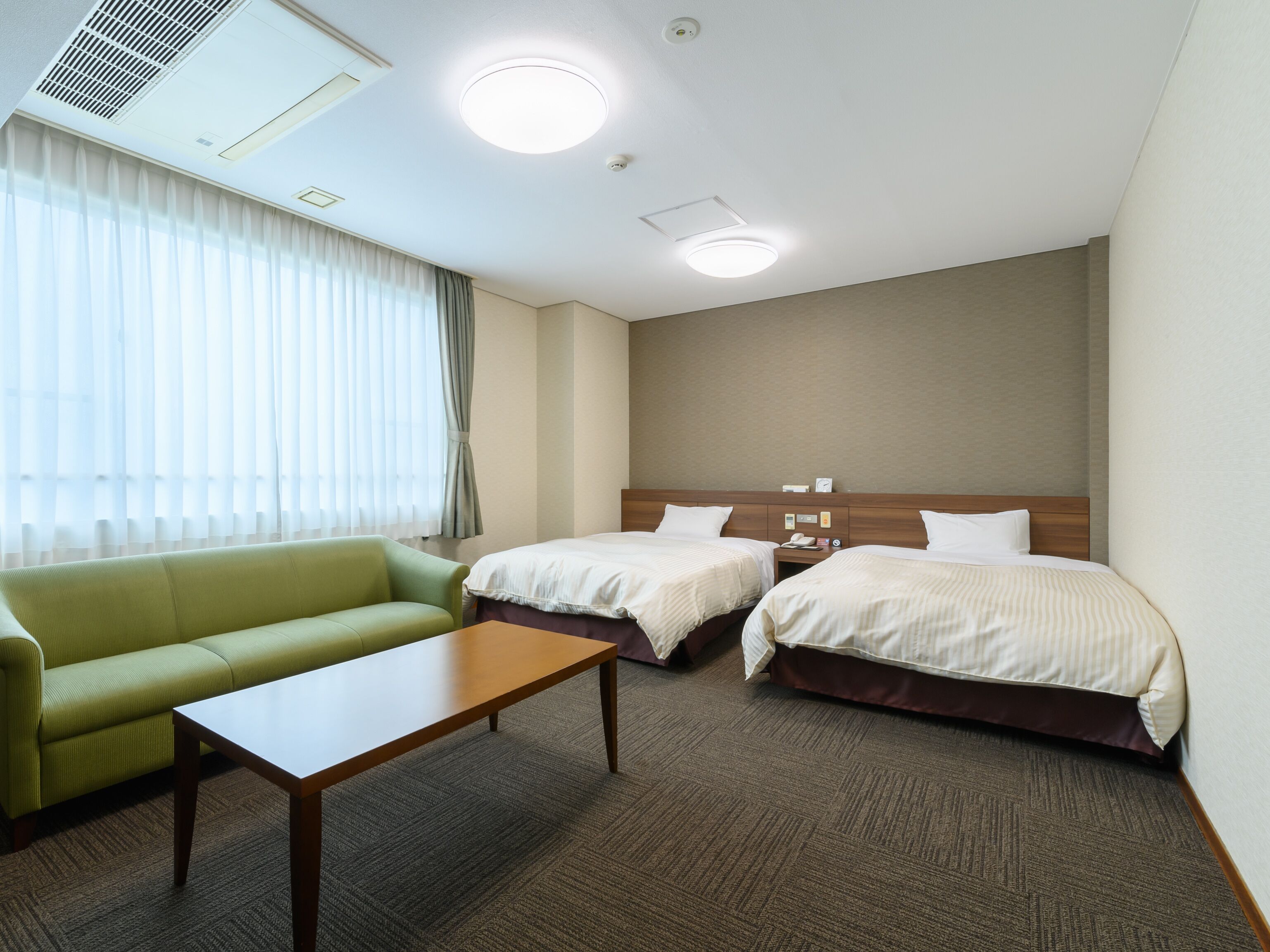A hotel room with two beds, a green sofa, a wooden table, and a large window with sheer curtains.