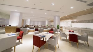 Zona pranzo moderna con tavoli bianchi e sedie rosse, un bancone centrale e un'area cucina sullo sfondo.