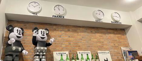 A hotel lobby with a brick wall, clocks labeled with city names, and a shelf with bottles and city-themed books.
