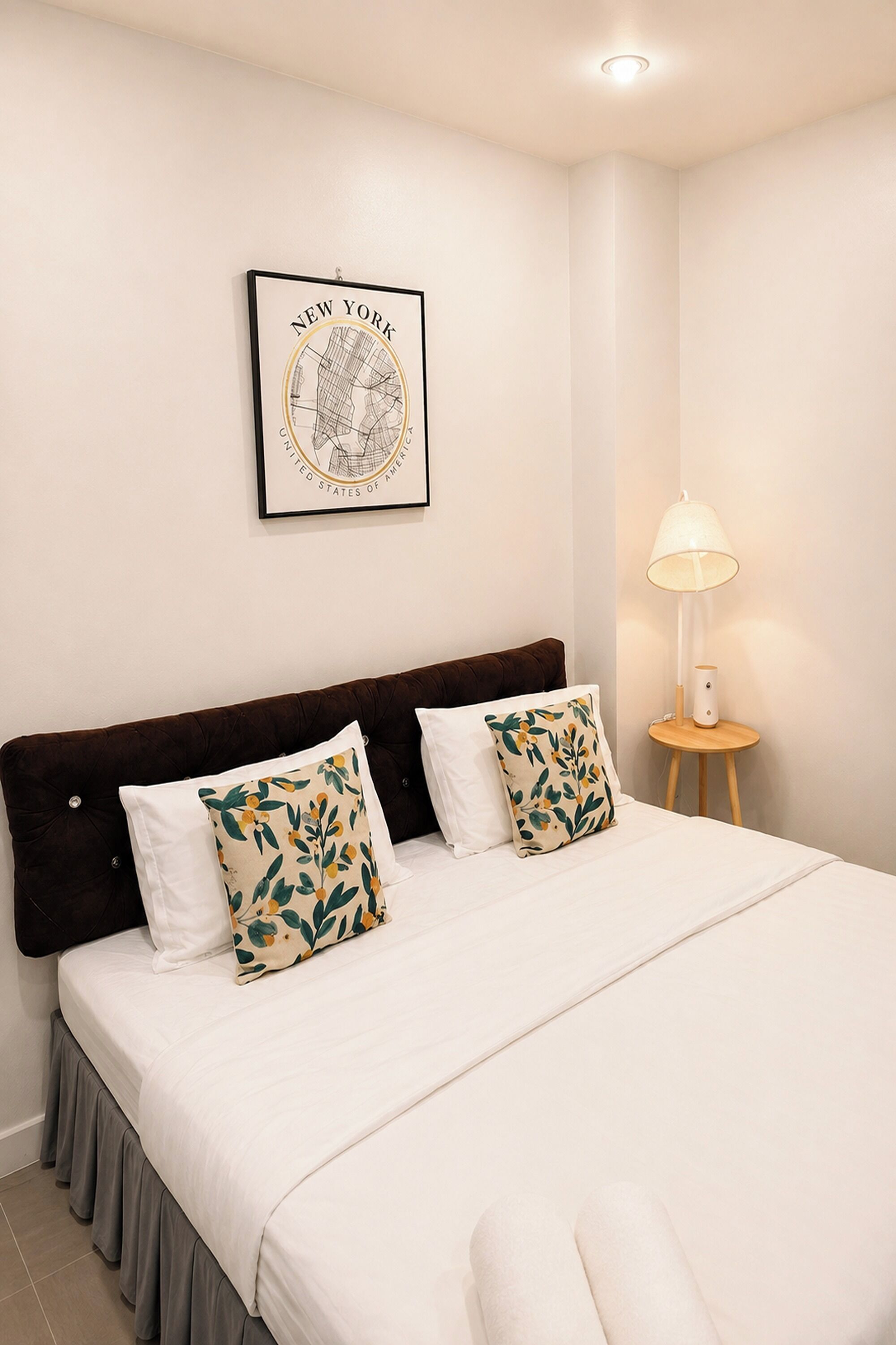 A neatly made bed with white bedding and two decorative pillows, a framed map of New York on the wall, and a bedside lamp on a small wooden table.