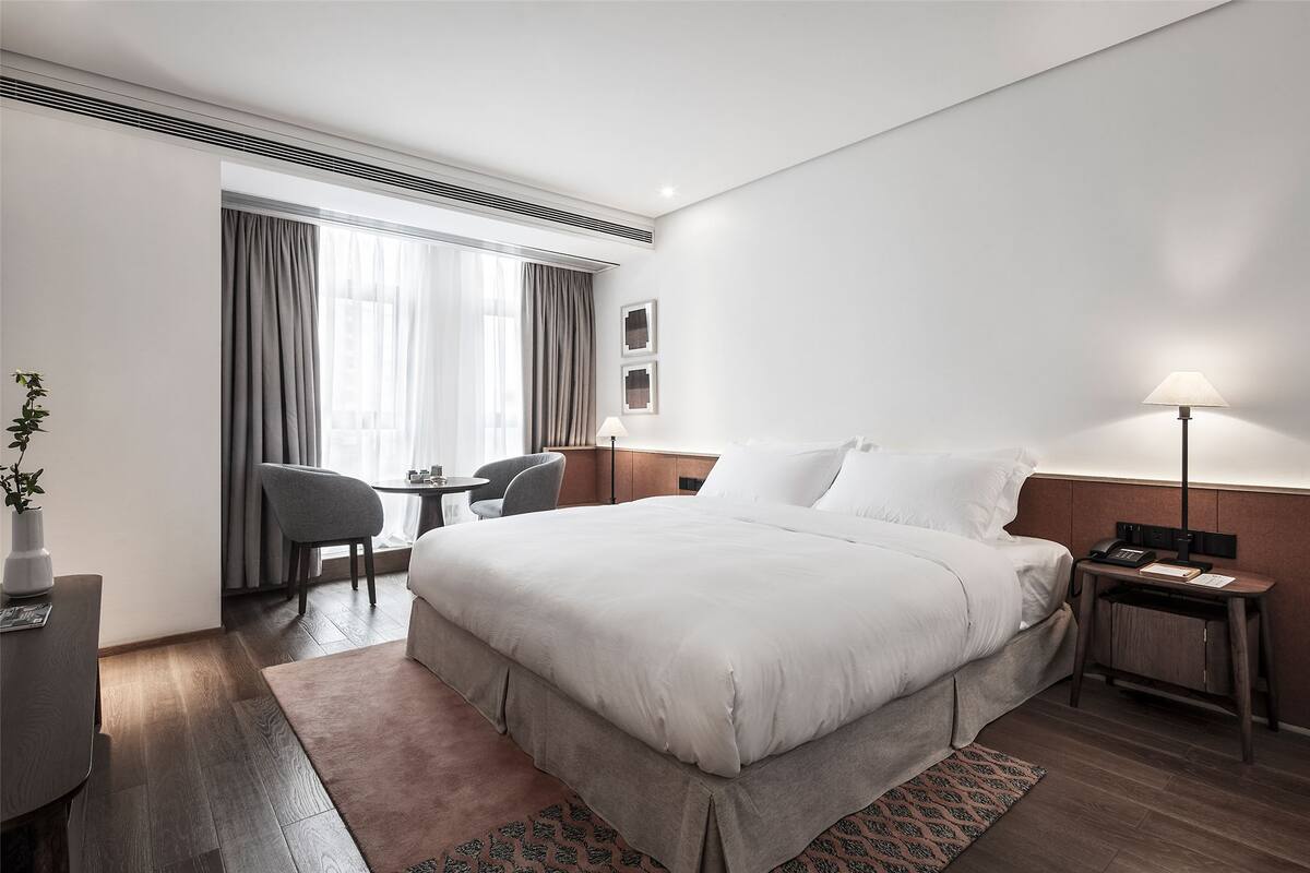 A hotel room with a large bed, a desk, a chair, a bedside table with a lamp, and a window with curtains.